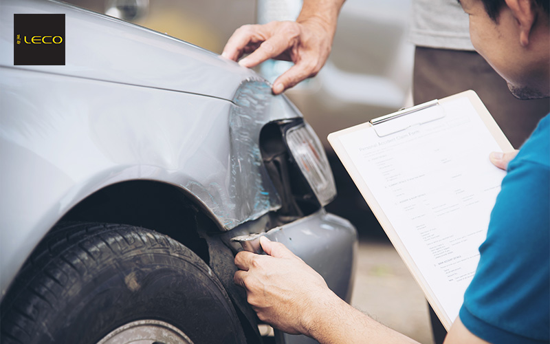 An insurance adjuster inspecting a dented silver car and holding a motor insurance claim form.