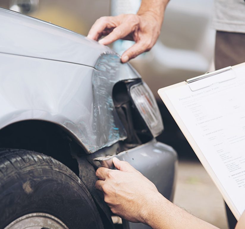 An insurance adjuster inspecting a dented silver car and holding a motor insurance claim form.