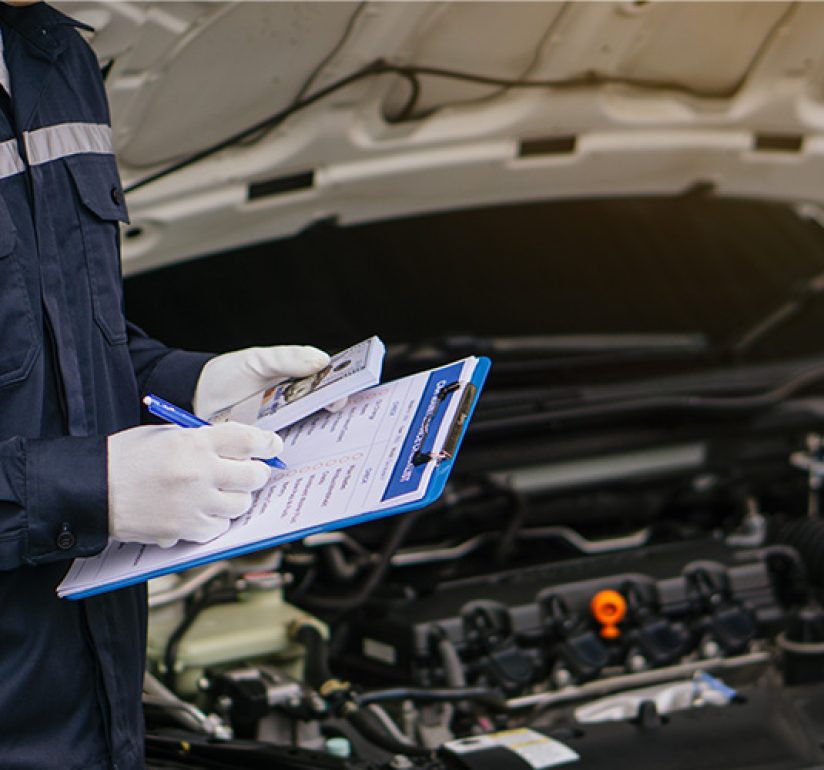 A car mechanic wearing gloves inspects an open engine bay while writing notes on a clipboard.