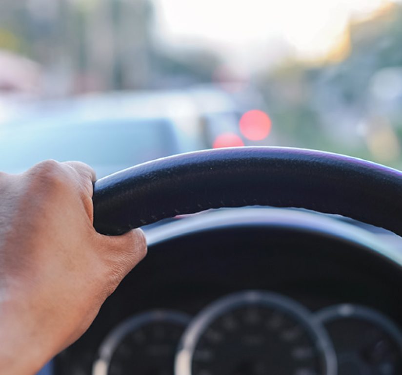 Driver’s view inside a car with hand on steering wheel, dashboard visible, and traffic ahead.